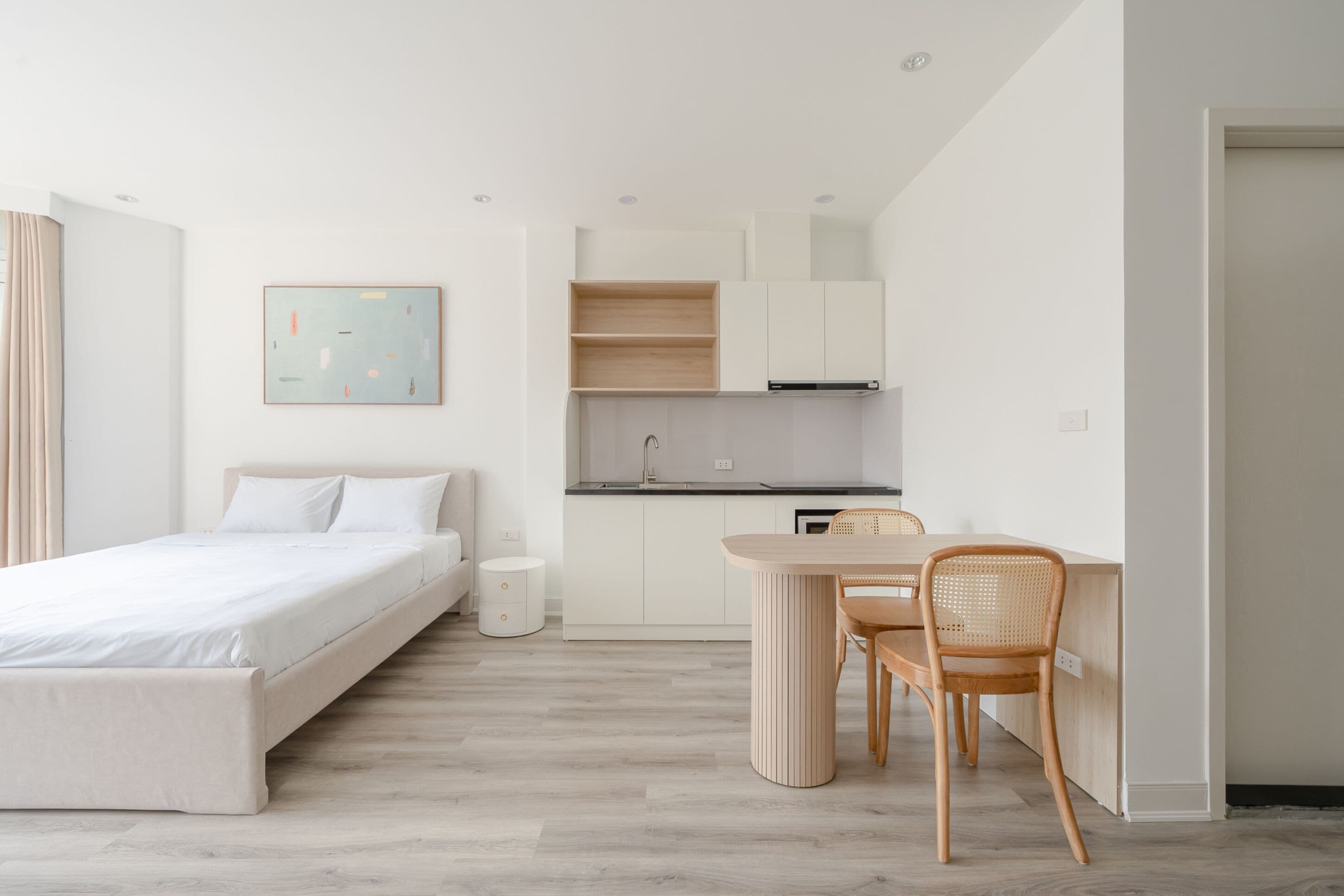 Un dormitorio moderno y minimalista con una cama individual, una mesita de noche pequeña y un velador. Junto al dormitorio hay una kitchenette compacta con una encimera y un horno empotrado.