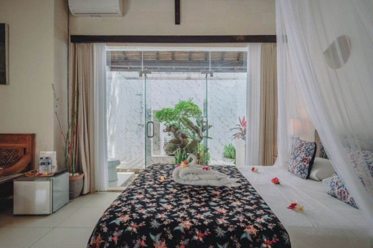 Habitación de hotel con una cama, una mesita con una planta y vistas a un jardín a través de un ventanal.