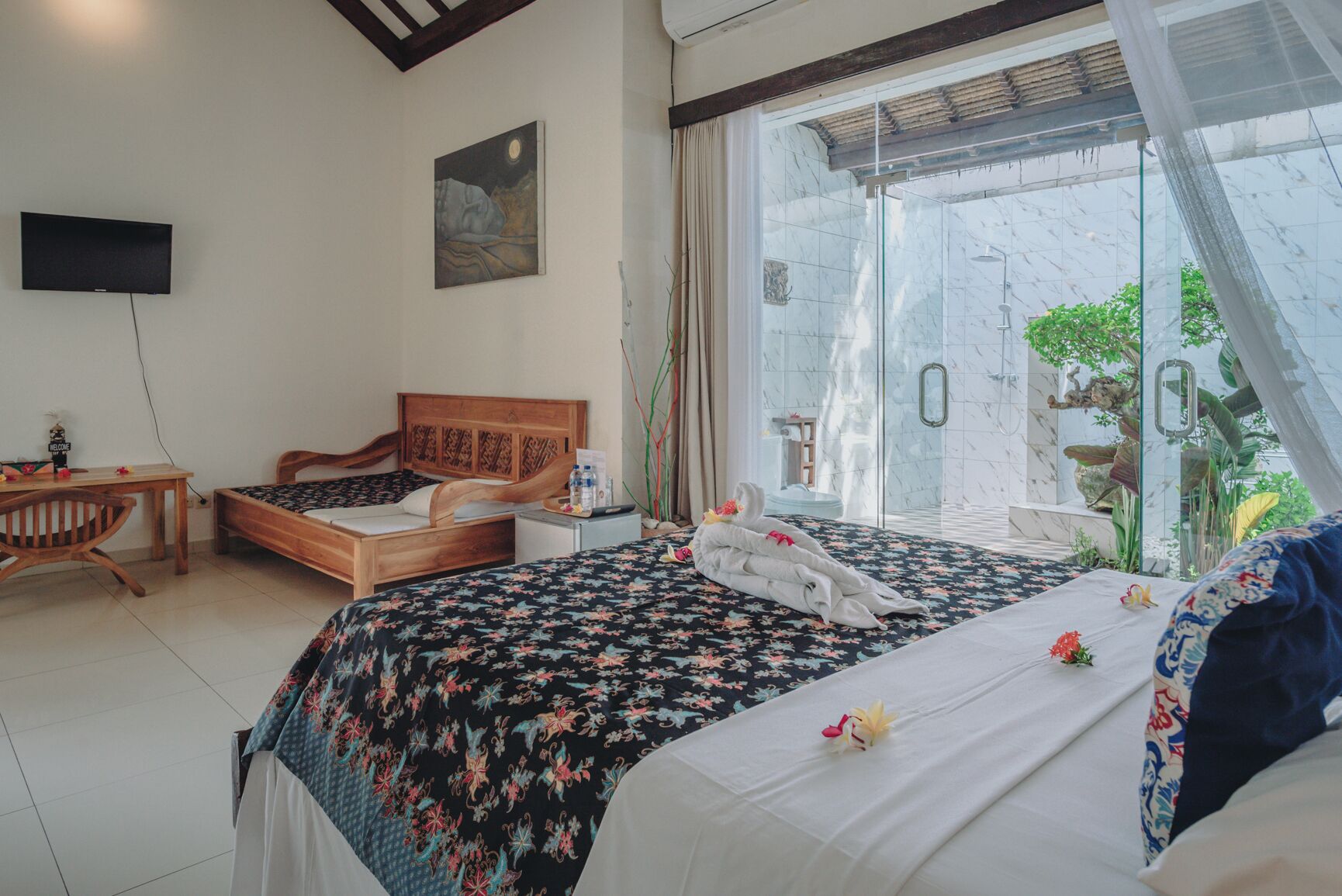 Un dormitorio con dos camas de madera, un escritorio, una silla, un televisor y una zona de ducha de vidrio con vista a la vegetación.