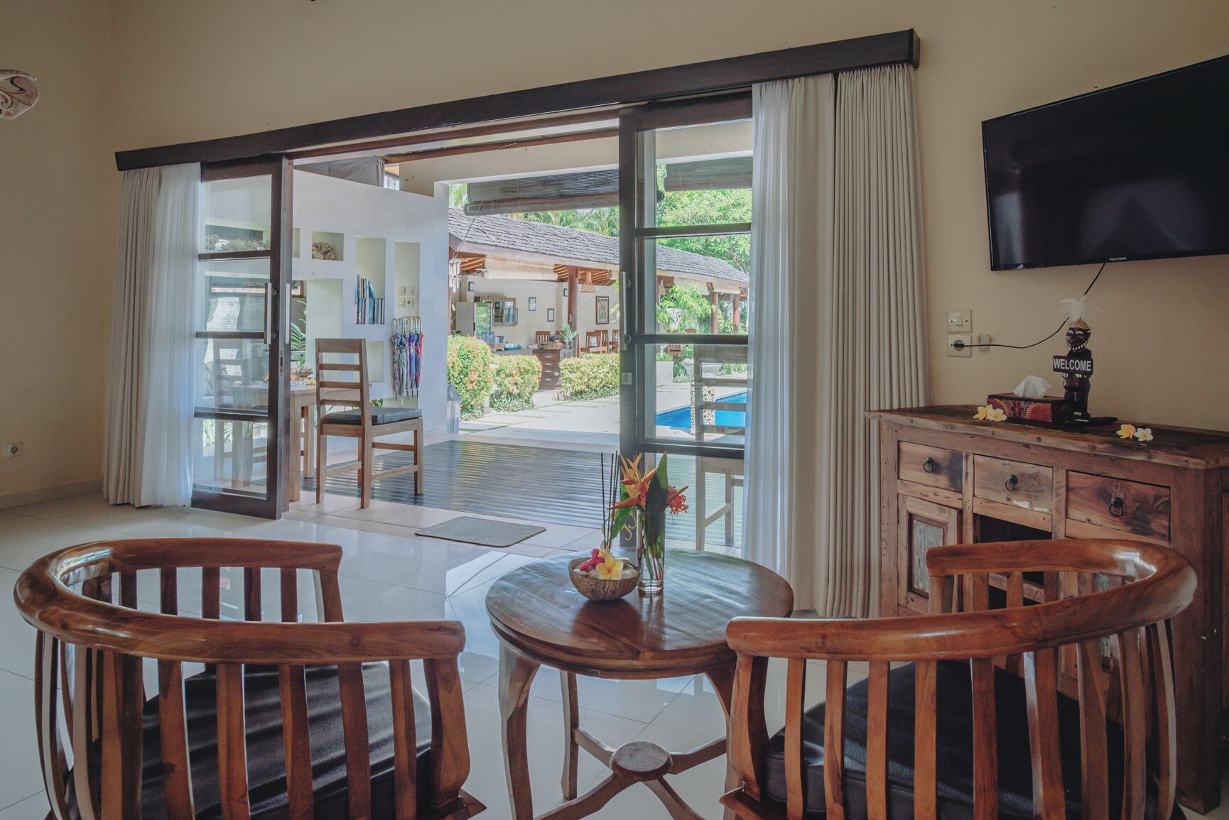 Una habitación con puerta de vidrio corredera que da a una zona de piscina, una mesa de madera con sillas, un televisor en la pared y un cartel de bienvenida.