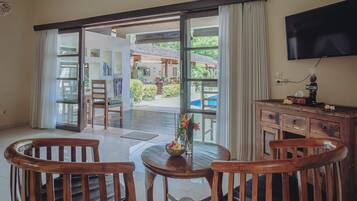 Una habitación con puerta de vidrio corredera que da a una zona de piscina, una mesa de madera con sillas, un televisor en la pared y un cartel de bienvenida.