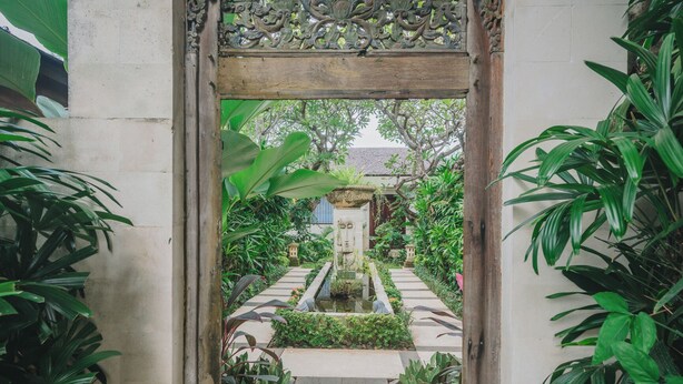 Um caminho de jardim leva a uma fonte central, cercada por vegetação exuberante e um arco de madeira decorativo.