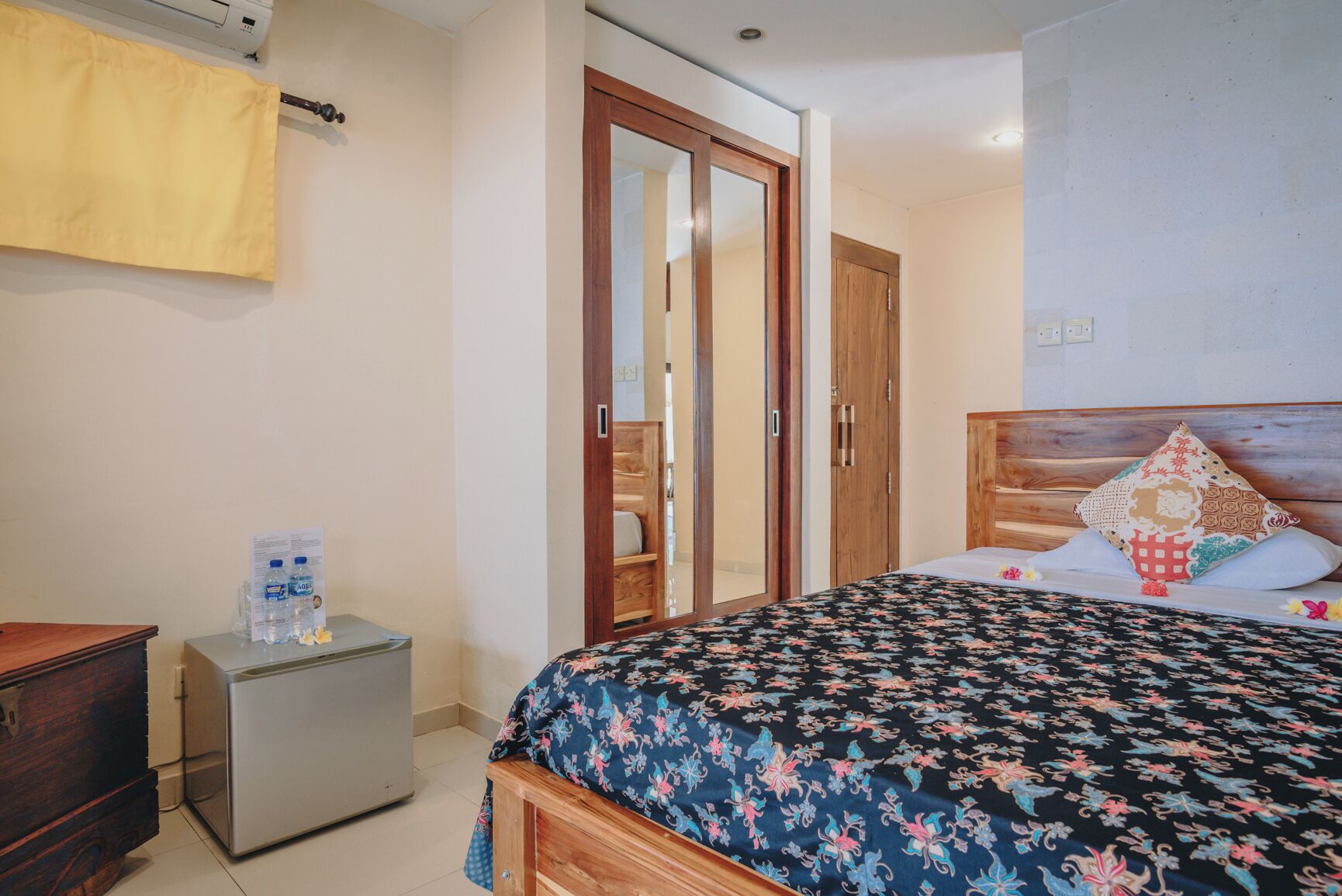 A bedroom with a bed, a wooden wardrobe, a small bedside table with water bottles, and a yellow curtain.