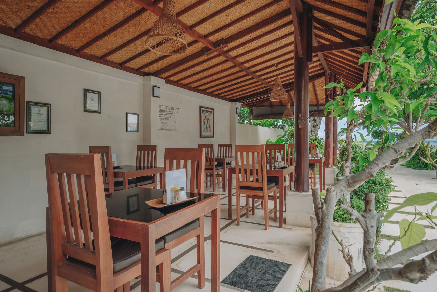 A covered outdoor dining area with wooden tables and chairs, a thatched roof, and a view of greenery.