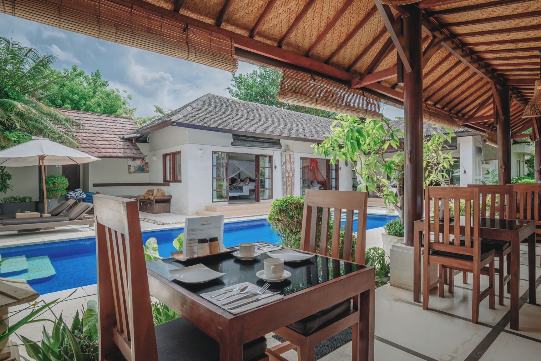 A poolside dining area with wooden furniture, a table set for two, and a view of a house with a tiled roof and a pool.