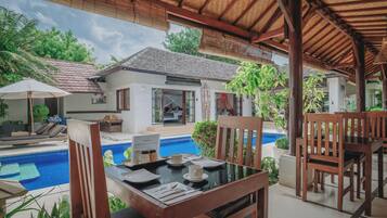 A poolside dining area with wooden furniture, a table set for two, and a view of a house with a tiled roof and a pool.