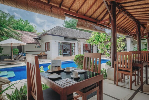 A poolside dining area with wooden furniture, a table set for two, and a view of a house with a tiled roof and a pool.