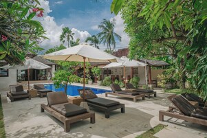 A poolside area with lounge chairs and umbrellas, surrounded by lush greenery and a clear sky.