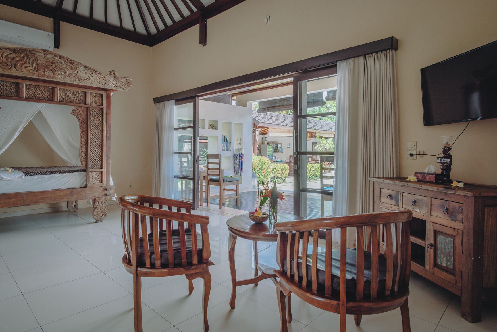 A room with a bed, two wooden chairs, a table with a vase, a cabinet, and a TV on the wall.