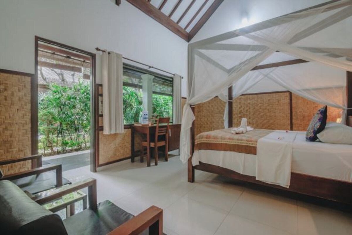 Un dormitorio con una cama, un escritorio y una silla. Se ve follaje verde a través de la ventana.