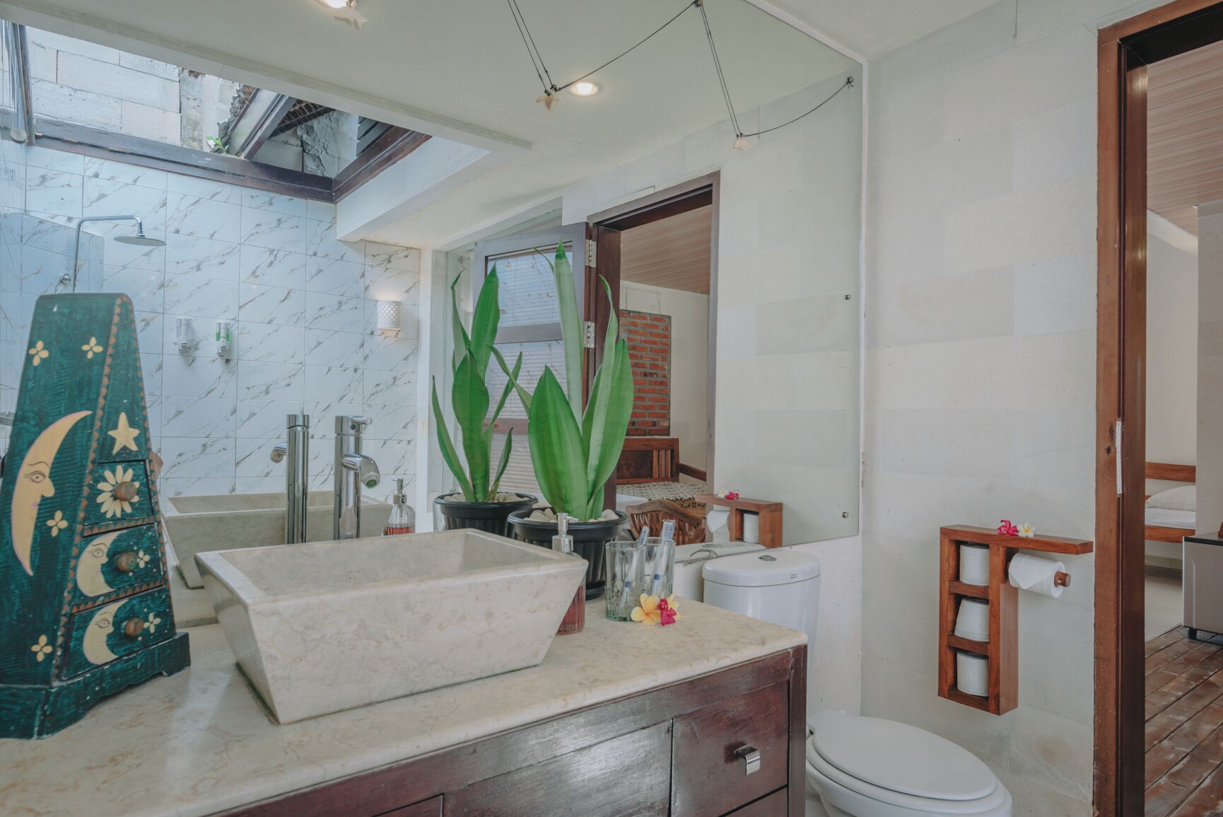 Ein Badezimmer mit einem großen Waschbecken, einer Toilette, einer Dusche und einem Spiegel. Auf der Ablage stehen Pflanzen und ein Holzregal mit Pflegeprodukten.