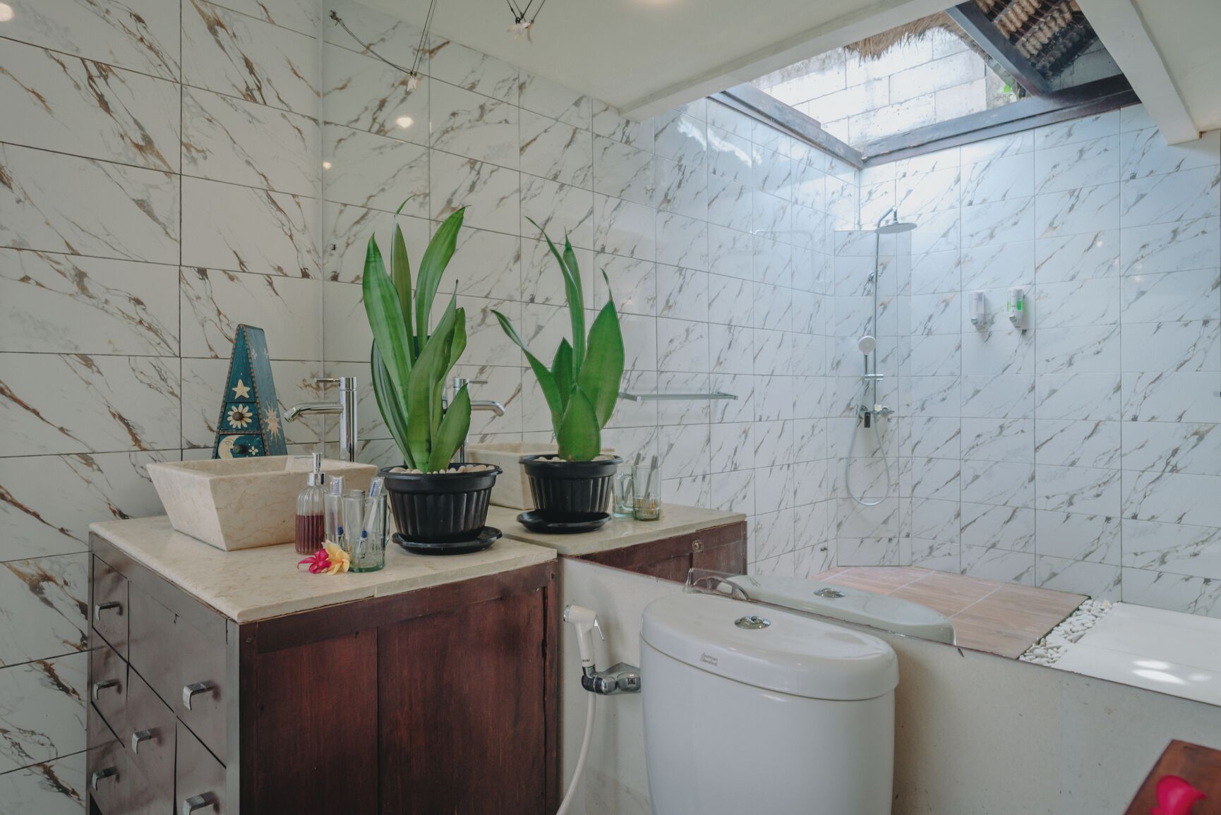 A bathroom with marble tiles, a wooden vanity, a toilet, a shower, and two potted plants.