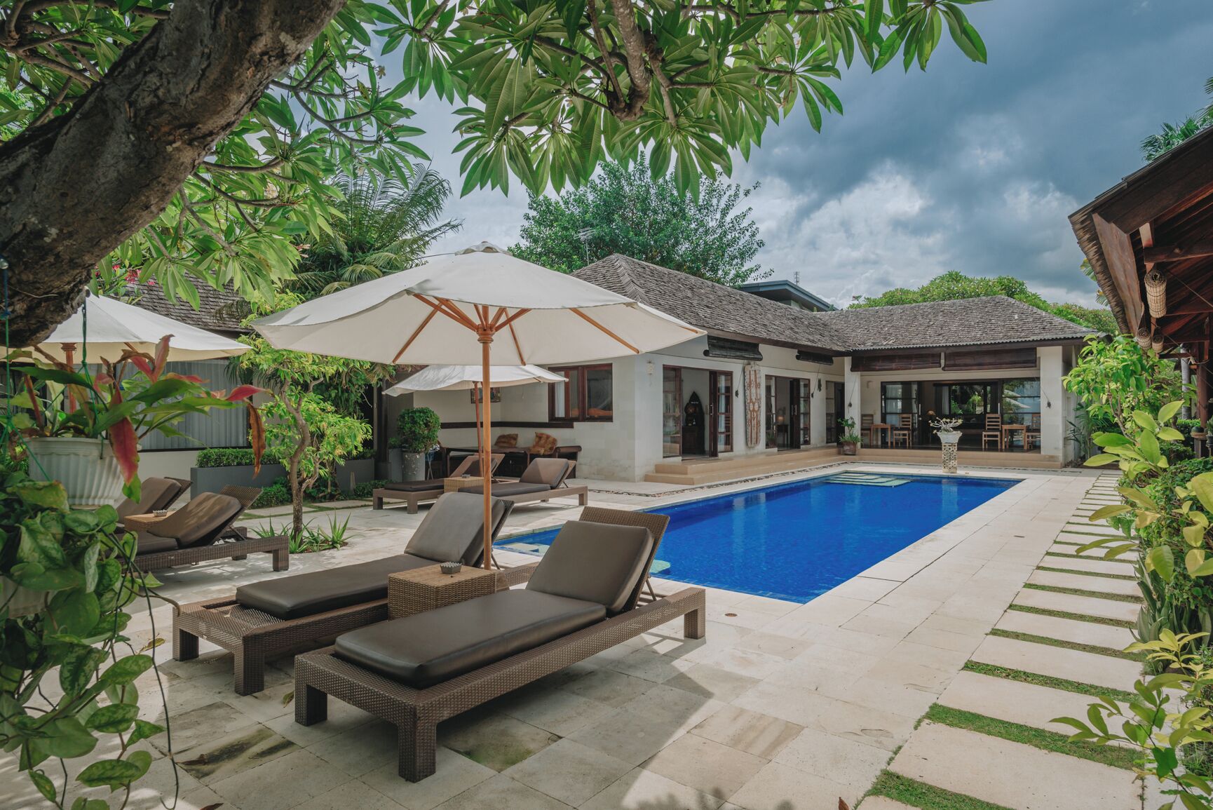 Un espace piscine agrémenté de chaises longues et de parasols, entouré d’une végétation luxuriante et d’une maison au toit recouvert de tuiles.