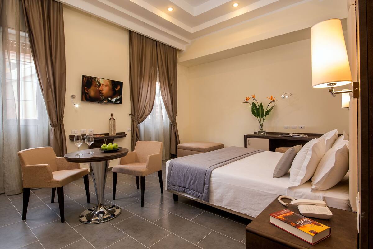 A hotel room with a bed, two chairs, a small round table with a vase and a bottle, a lamp, and a book on the bedside table.