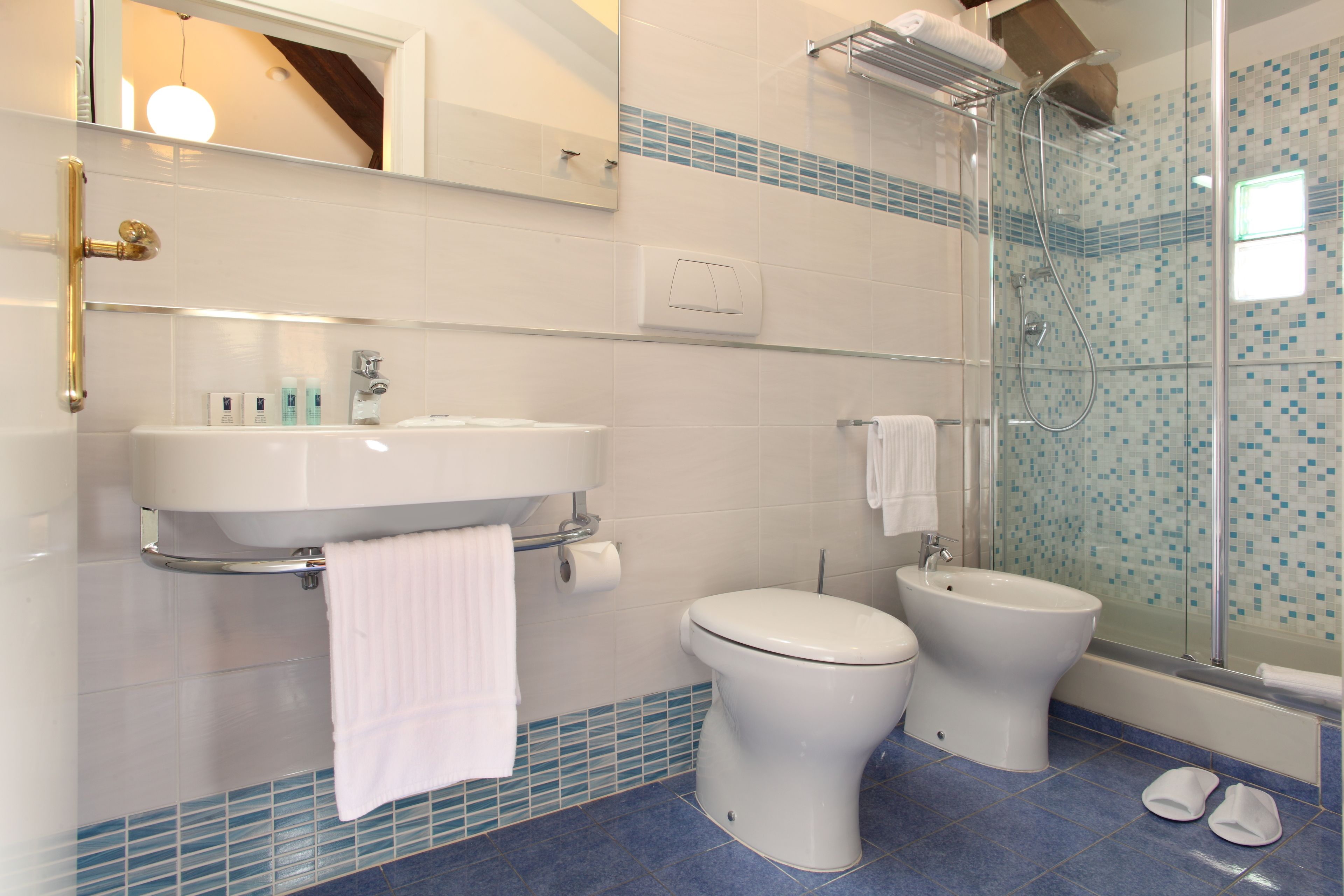 Une salle de bain avec un lavabo blanc, deux toilettes, une douche en verre et un sol carrelé de bleu.