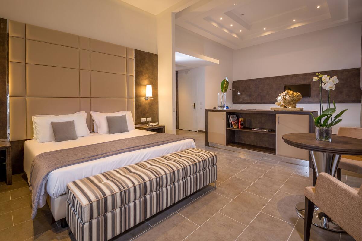 A modern hotel room with a large bed, a bedside table, a bench, a dining area with a table and chairs, and a kitchen area with a counter and decorative items.
