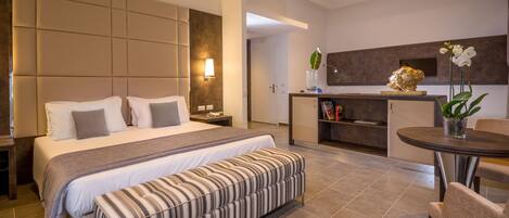 Una habitación de hotel moderna con una cama grande, una mesita de noche, un banco, un área de comedor con mesa y sillas, y una cocina con mostrador y detalles decorativos.