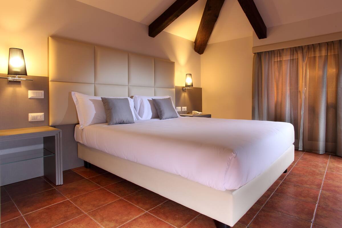 A modern hotel room with a large bed, two bedside tables, a nightstand, and a window with curtains.