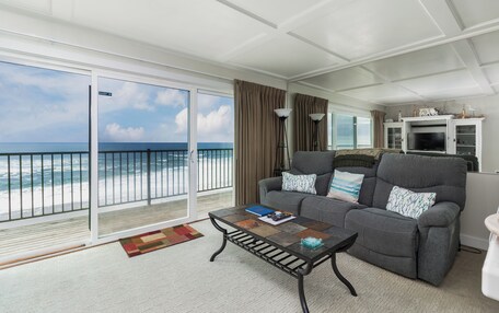 A living room with a balcony overlooking the ocean, a gray sofa, a coffee table, and a television set.