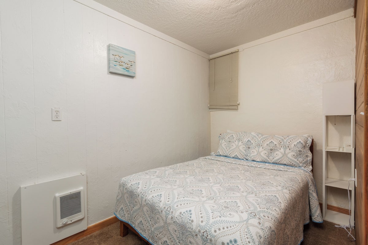 A small, neatly arranged bedroom with a patterned bedspread, a wall-mounted heater, and a window with blinds.