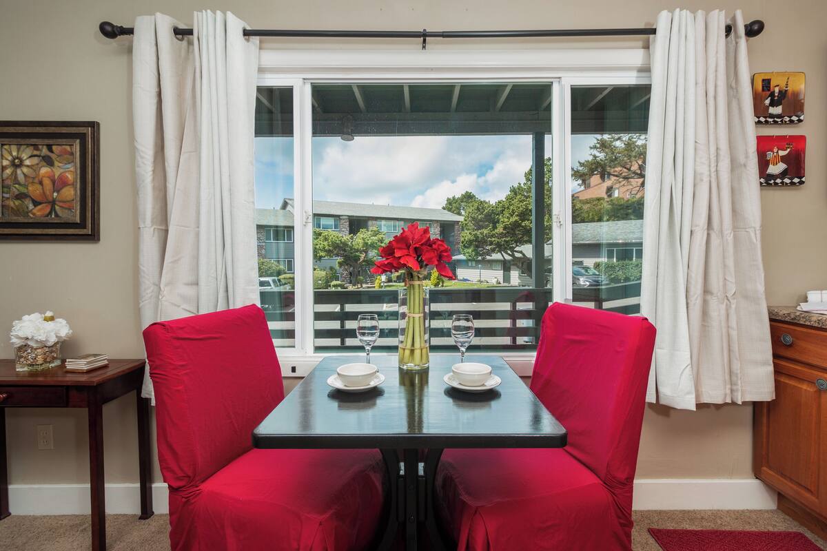 A dining area with a table set for two, red upholstered chairs, a vase with red flowers, and a view of a balcony with a railing and greenery.