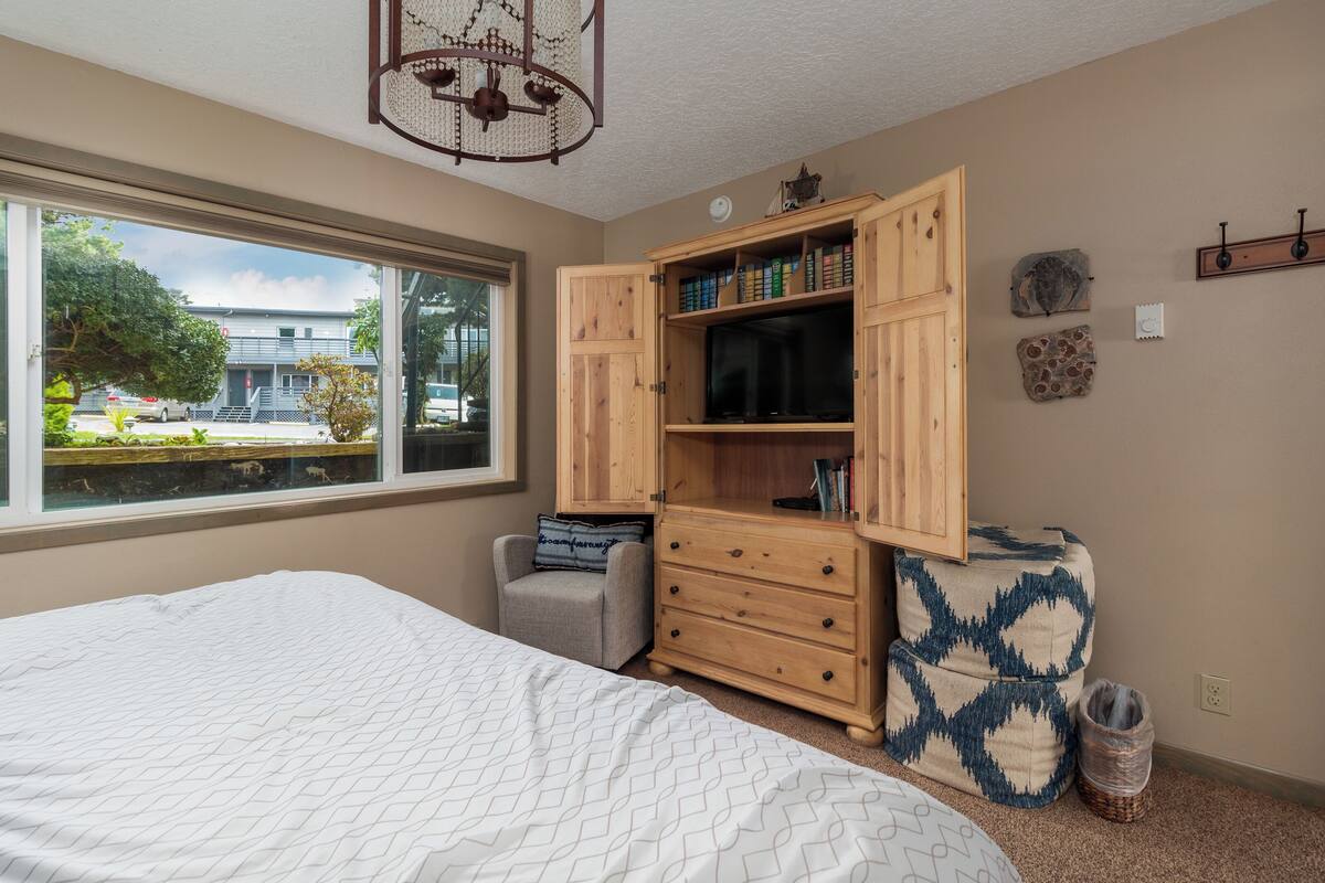 A bedroom with a large bed, a wooden wardrobe, a small chair, and a window with a view of a building and trees.
