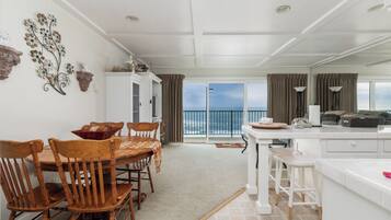 A spacious room with a dining table and chairs, a kitchen area, and a large sliding glass door leading to a balcony with a view of the ocean.
