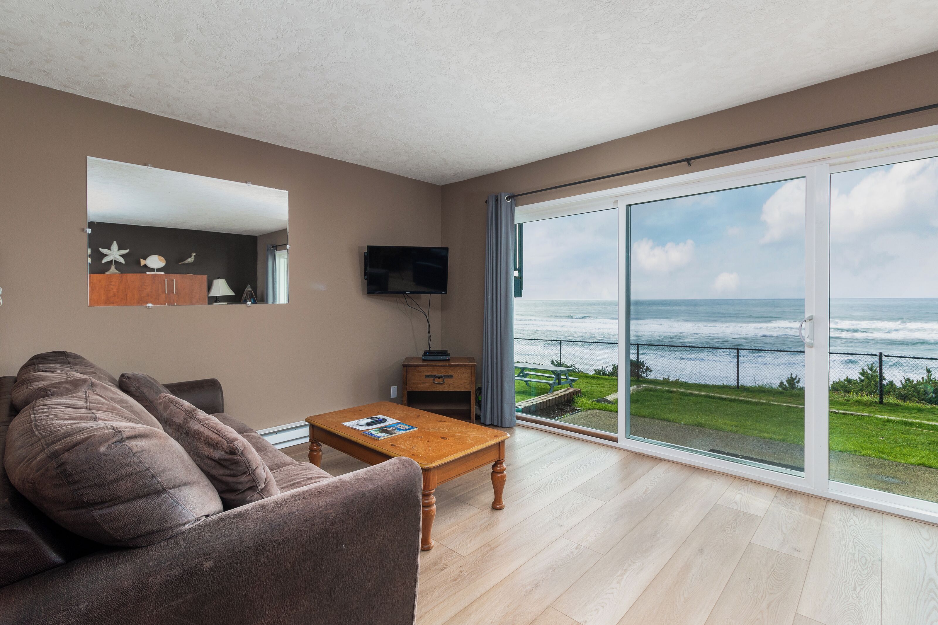 A living room with a sofa, a wooden coffee table, a flat-screen TV, and a large sliding glass door leading to a balcony with a view of the ocean.