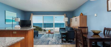 A room with a kitchen area, dining table, and a large window with a view of the ocean.