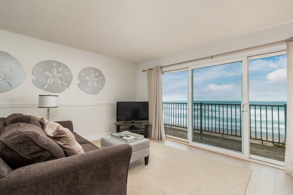 A living room with a sofa, a TV, and a view of the ocean through a sliding glass door.