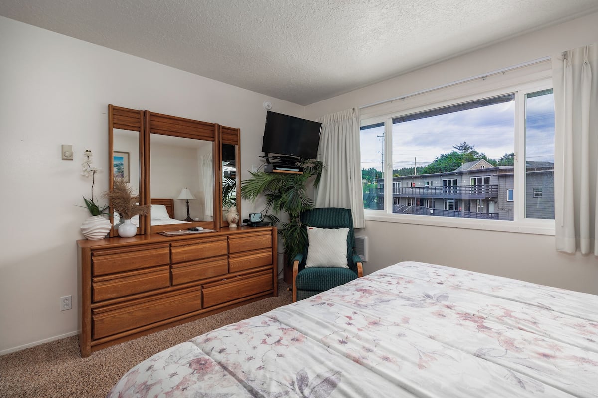 A bedroom with a wooden dresser, a bed with a floral bedspread, a green armchair, a large mirror, a flat-screen TV, and a window with a view of buildings.