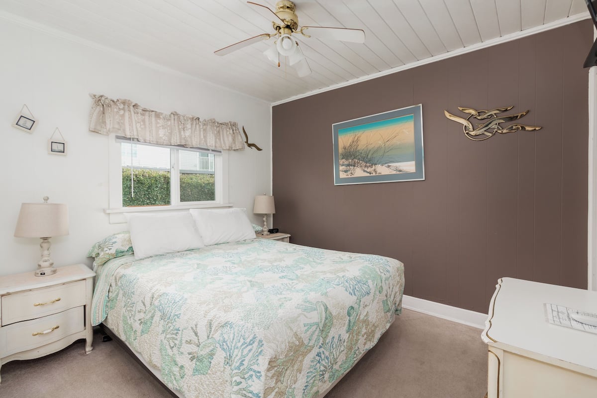 A bedroom with a bed, a nightstand, a dresser, a ceiling fan, a window with curtains, a wall-mounted picture, and wall art.