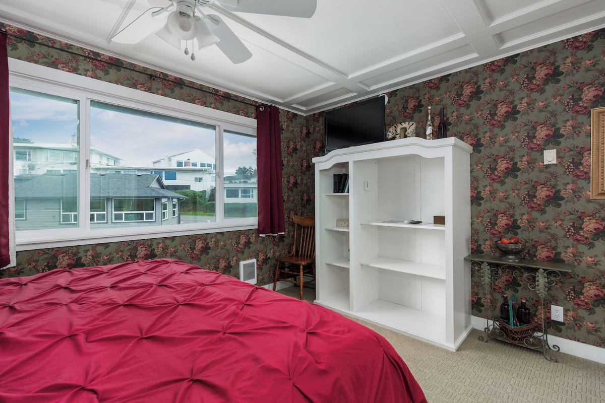 A bedroom with a large bed, a wardrobe, a television, and a window with a view of houses.