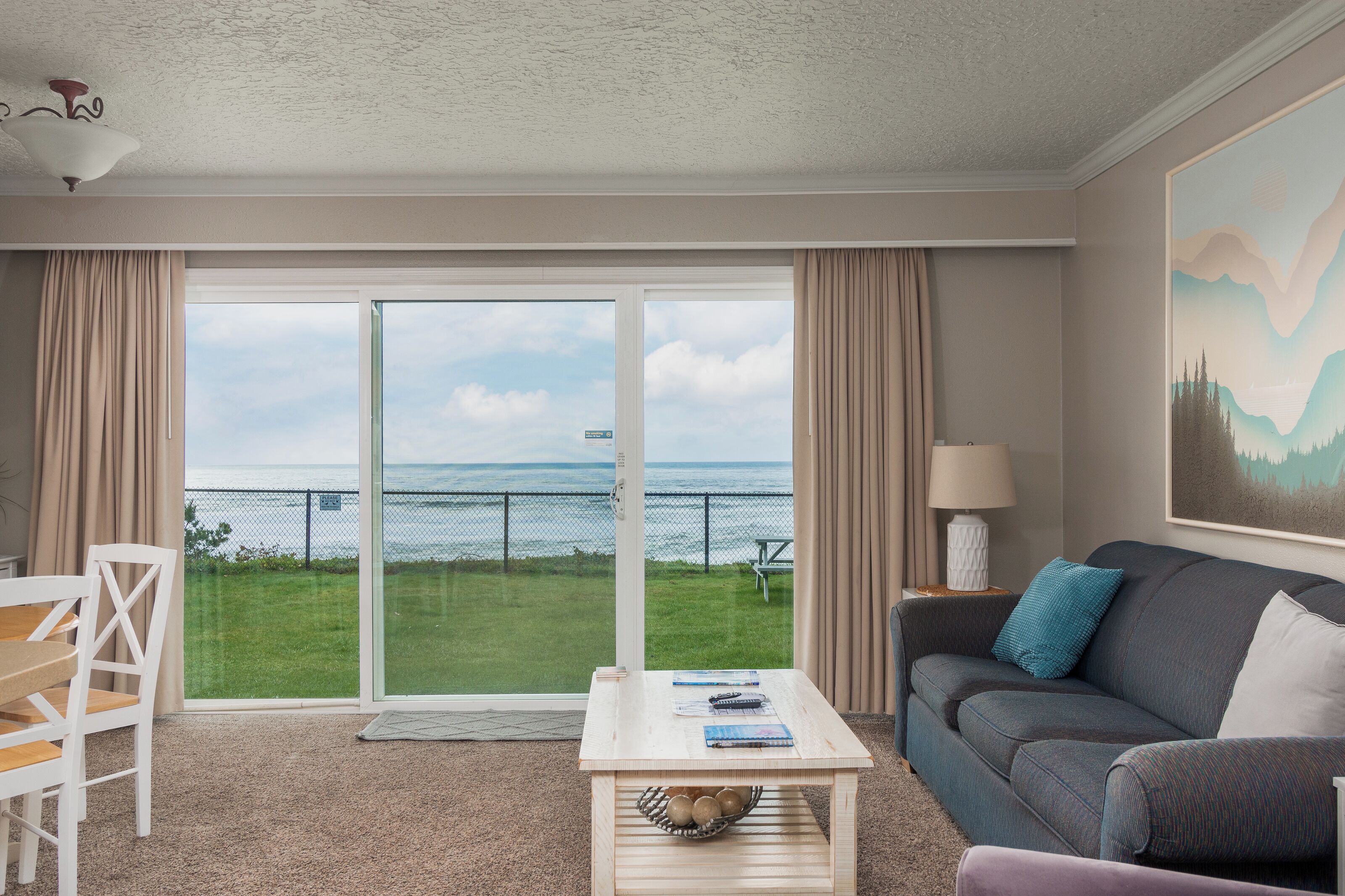 A room with a large sliding glass door leading to a grassy area with a view of the sea. There is a white table with a basket, a grey sofa with a blue pillow, and a lamp on a side table.