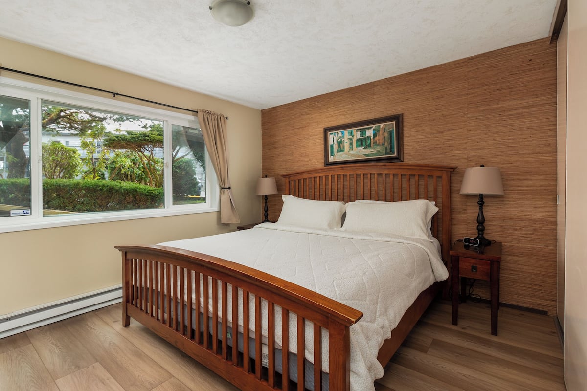 A bedroom with a wooden bed, a nightstand, a lamp, a window with a view of greenery, and a painting on the wall.