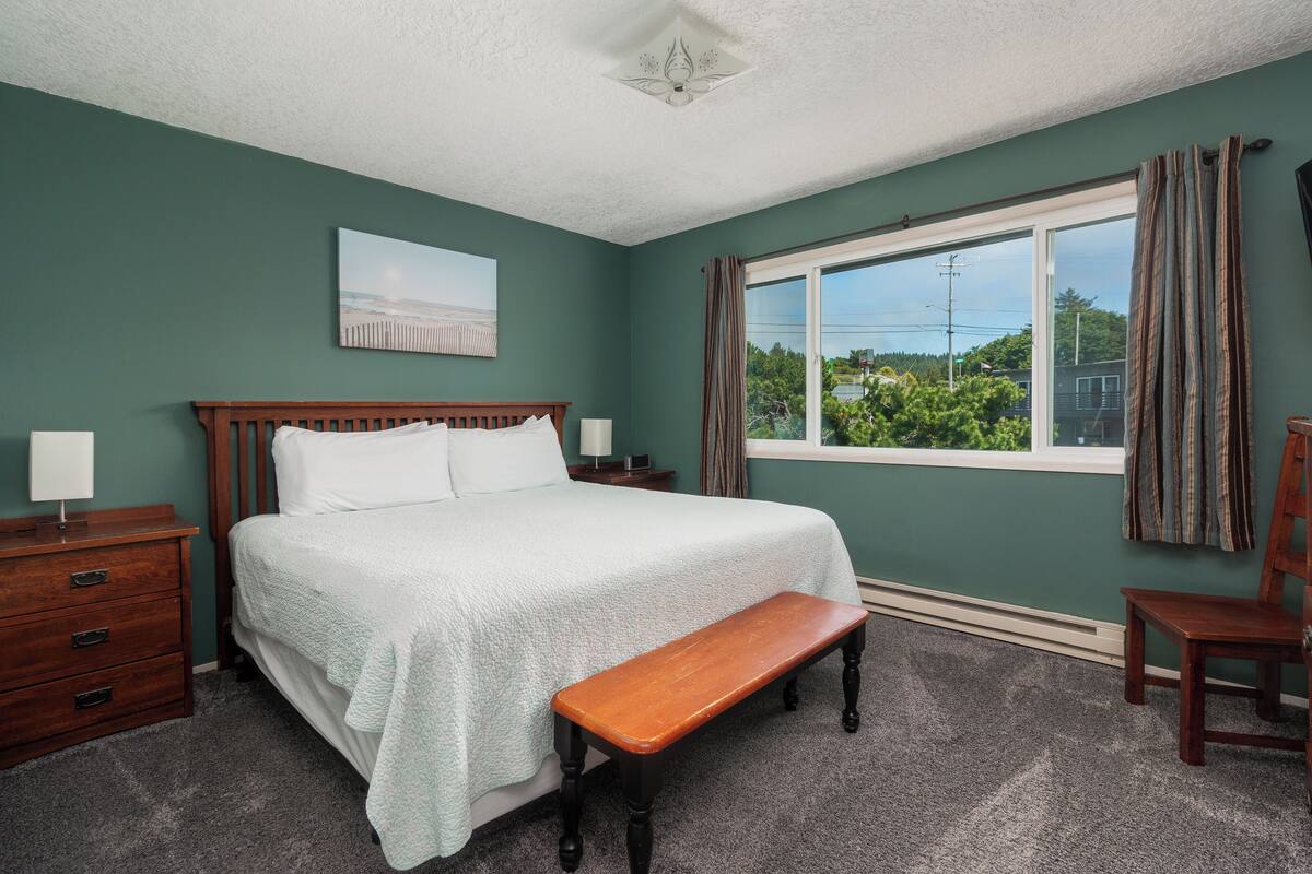 A bedroom with a large bed, a wooden bench, a nightstand, a dresser, and a window with curtains.
