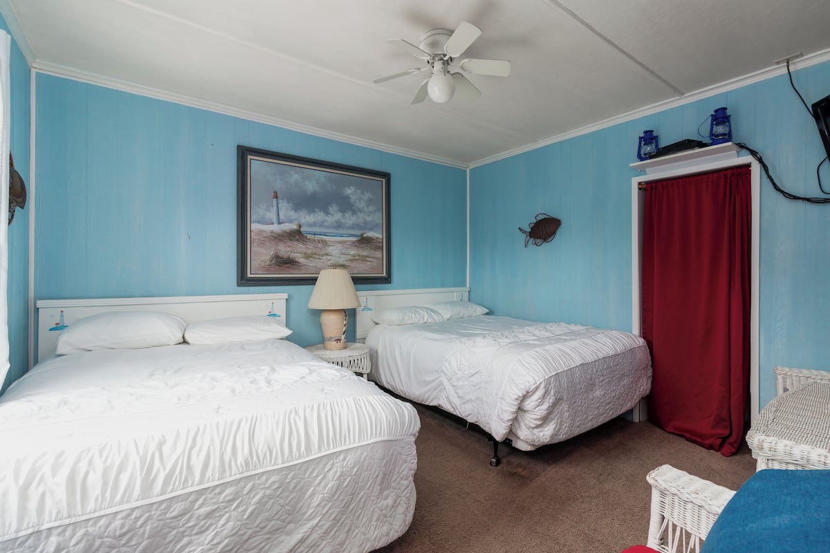 A bedroom with two beds, a ceiling fan, a painting, a red curtain, and a small table with a lamp.