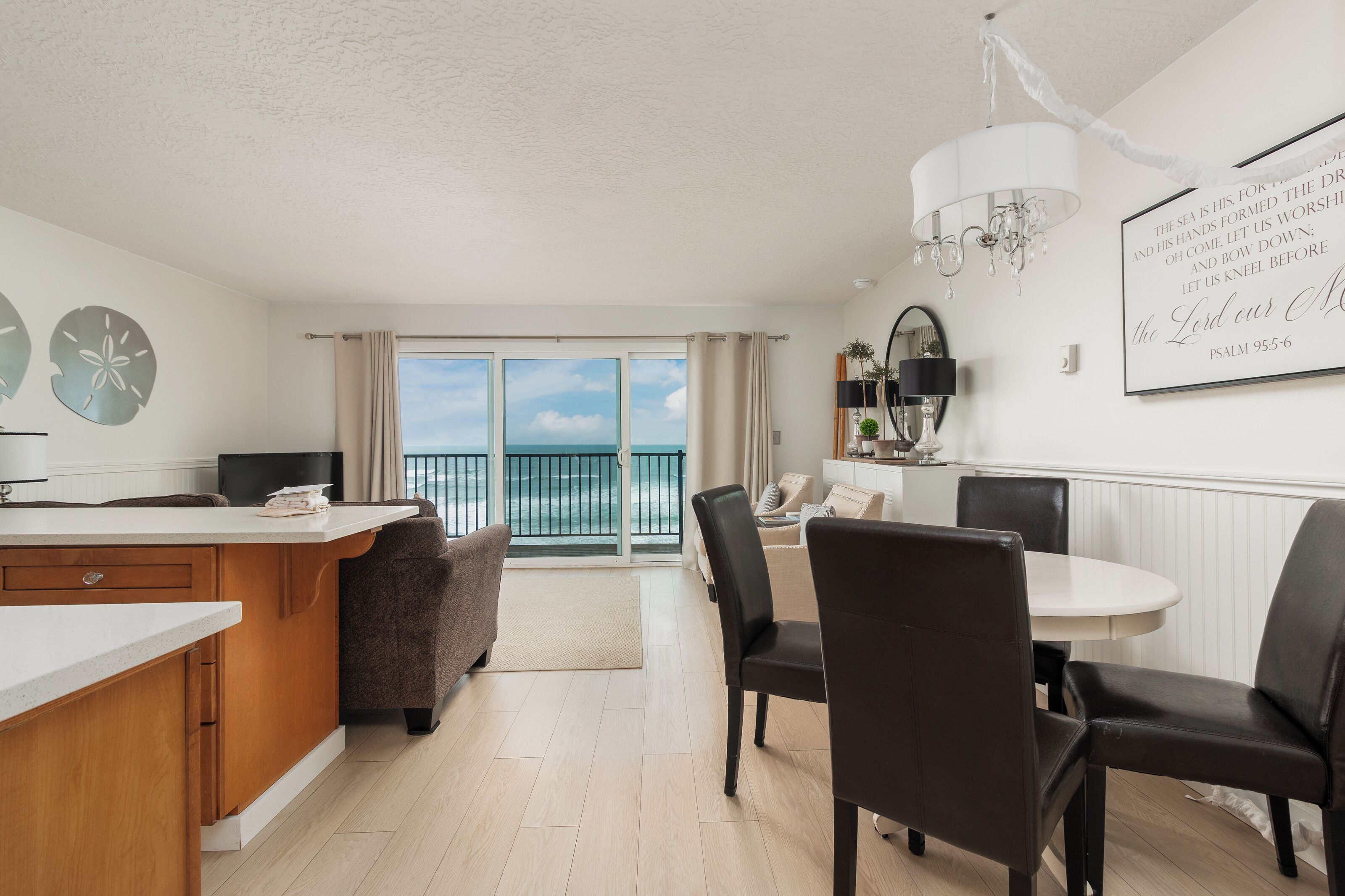 A modern kitchen with a large island, dining area with chairs, a chandelier, and a view of the sea.