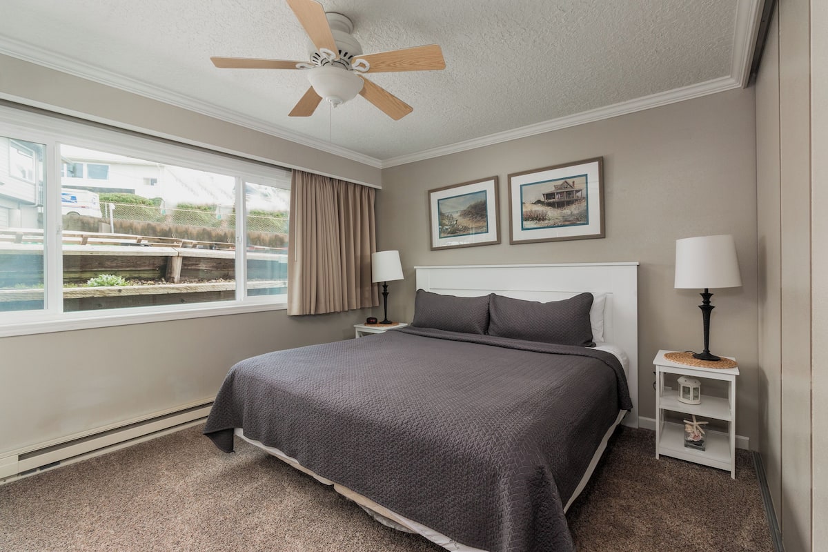 A bedroom with a bed, two bedside tables, a ceiling fan, a window with curtains, and two framed pictures on the wall.