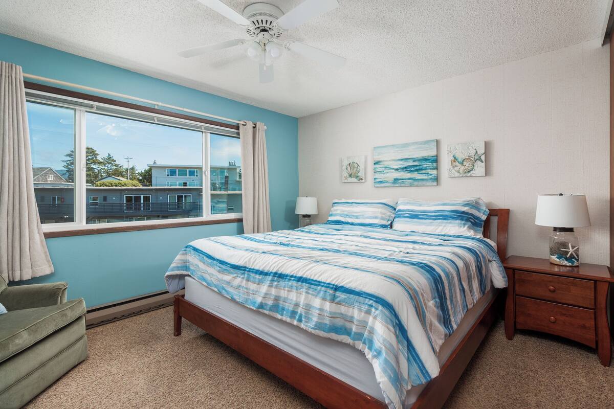 A bedroom with a large bed, a nightstand, a window with curtains, and artwork on the wall.