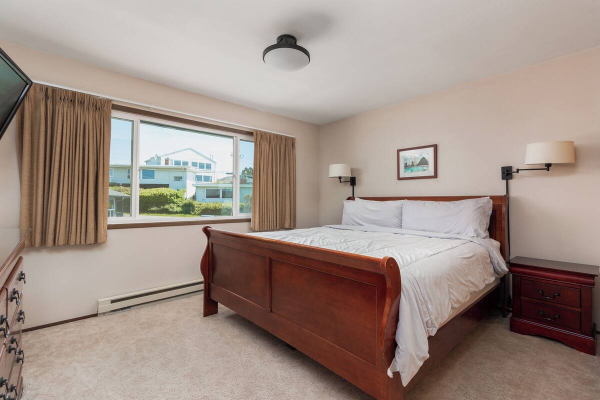 A bedroom with a wooden bed, a nightstand, a lamp, a window with curtains, and a picture on the wall.