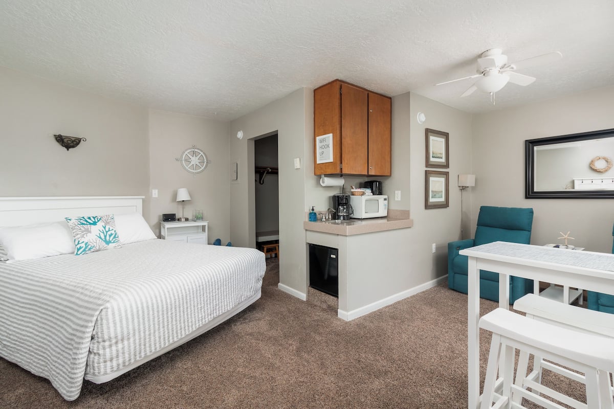 A bedroom with a bed, a desk, a chair, a kitchenette, and a ceiling fan.