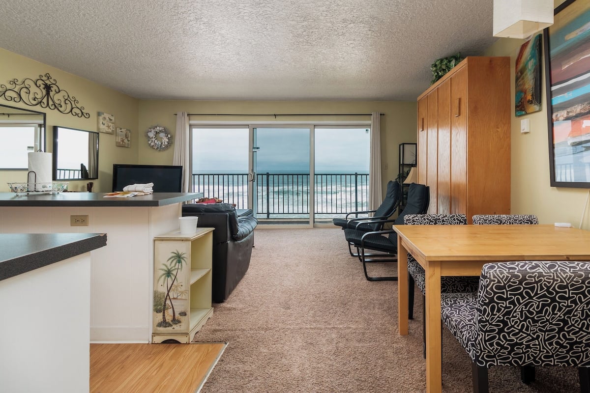 A living room with a view of the ocean, a dining area with patterned chairs, a wooden table, and a kitchen area with a counter and a decorative shelf.