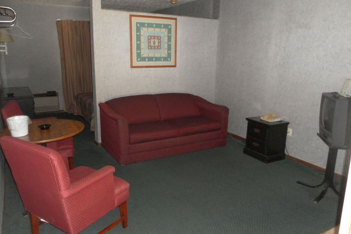 Um quarto pequeno e mobiliado com um sofá vermelho, uma cadeira de madeira, uma mesinha, uma televisão e um armário.