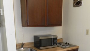 A compact kitchen with wooden cabinets, a stainless steel microwave, a small refrigerator, and a trash bin. - Tidewater Motel (Hayes)