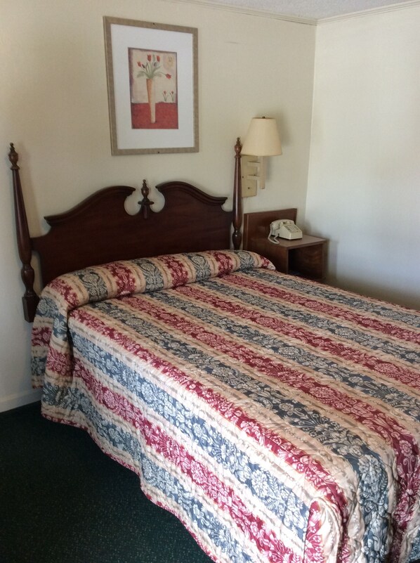 A neatly made bed with a floral-patterned bedspread, a wooden headboard, a bedside lamp, and a framed picture on the wall.
