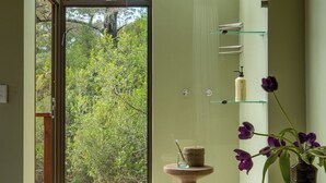 A modern bathroom with a glass shower enclosure, a wooden stool, a vase with flowers, and a view of greenery outside. - Tamodi Lodge and Stables (Keurboomstrand)