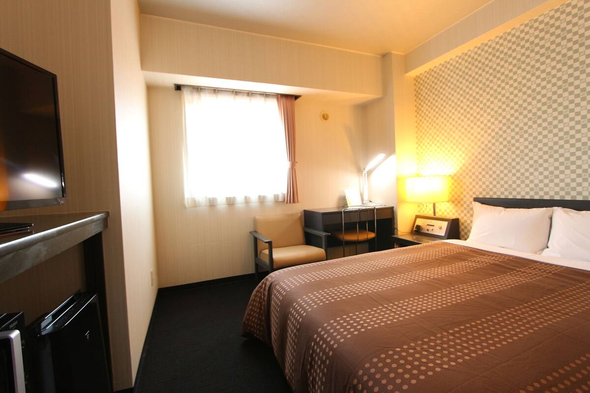 Una habitación de hotel con cama, escritorio con silla, televisor y ventana con cortinas.