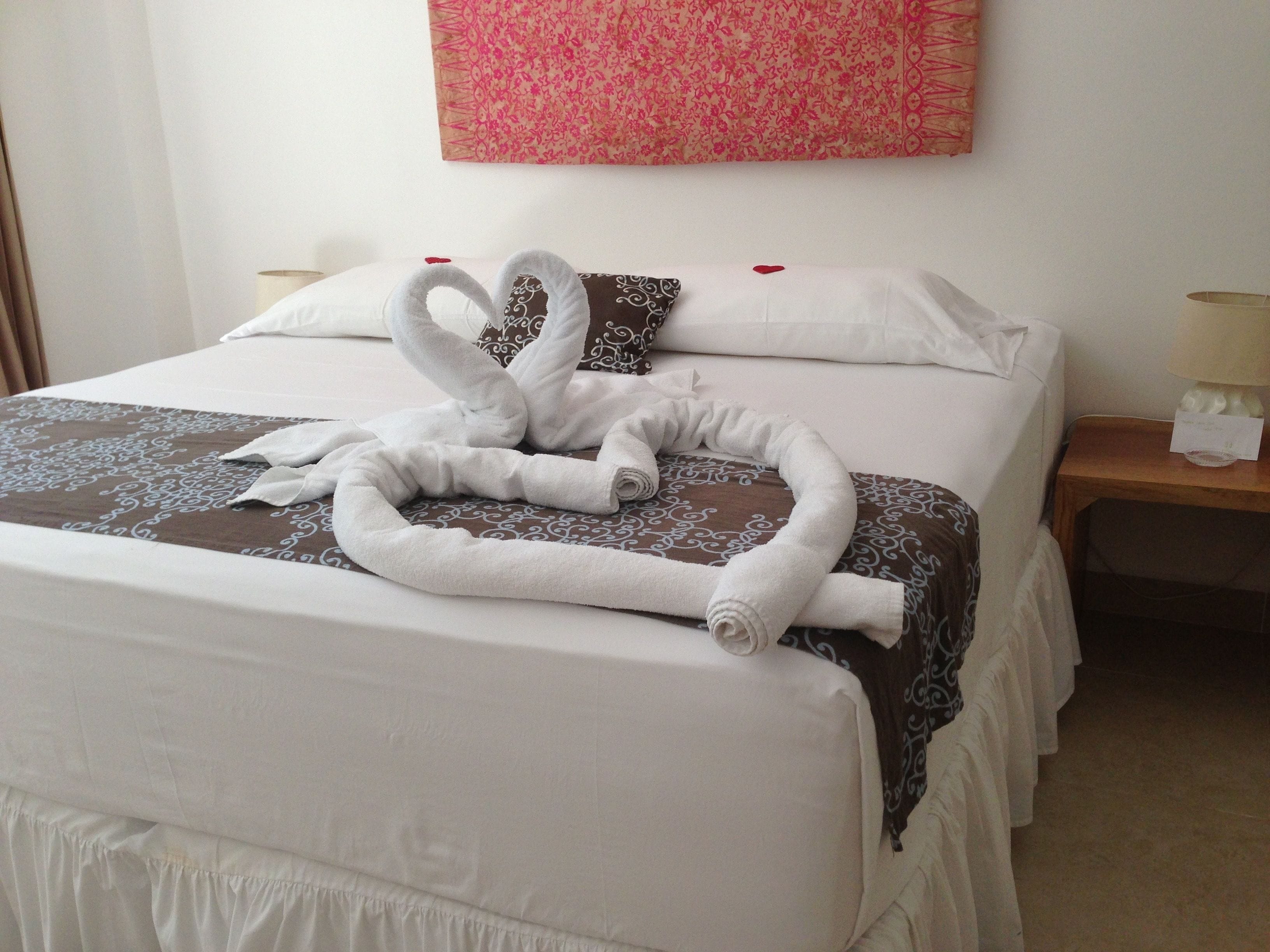 A neatly made bed with a towel arranged in a heart shape, a decorative pillow, and a pink wall hanging.