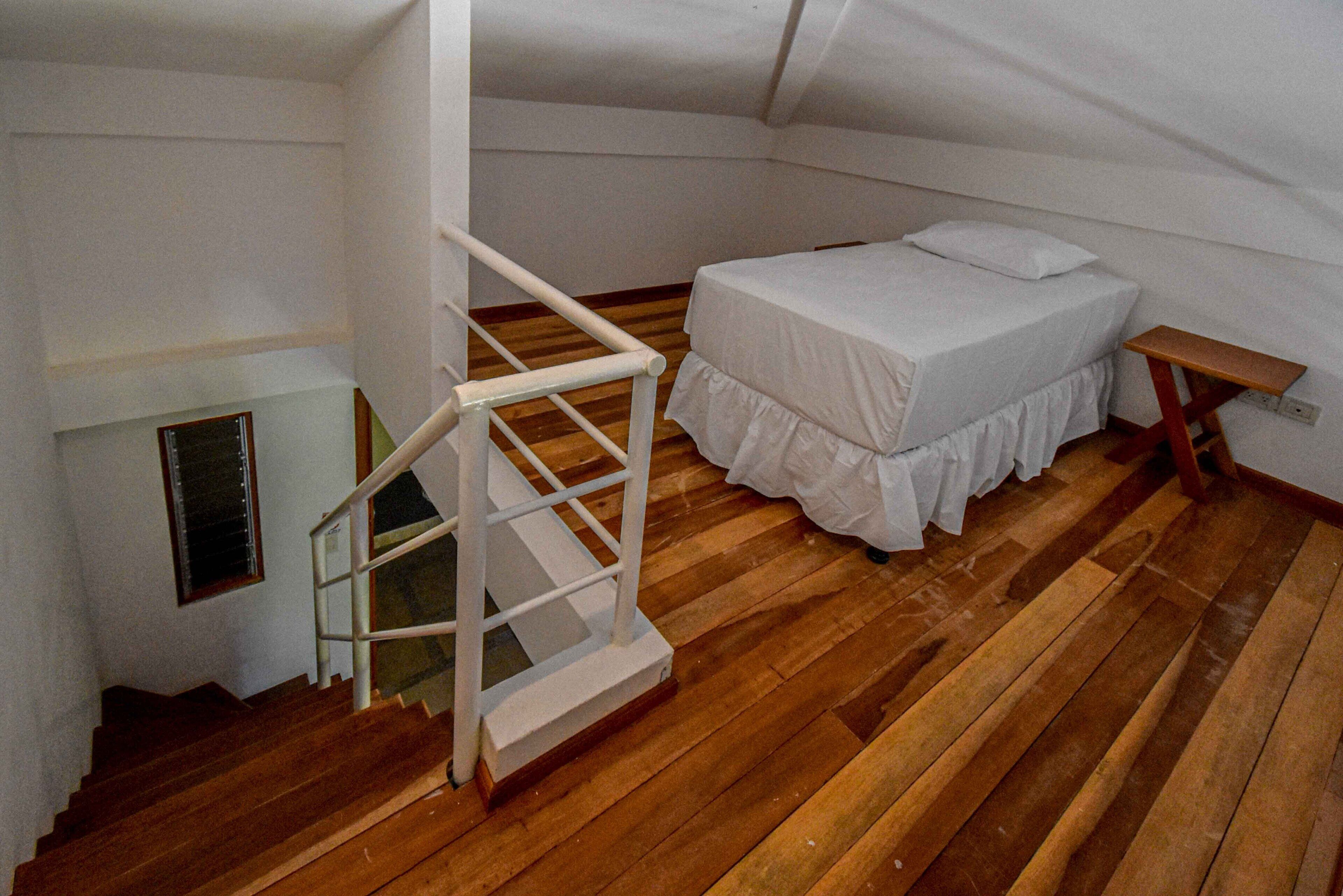 Un dormitorio con piso de madera, una cama blanca con cubrecama blanco, una mesita de noche blanca y una escalera blanca.